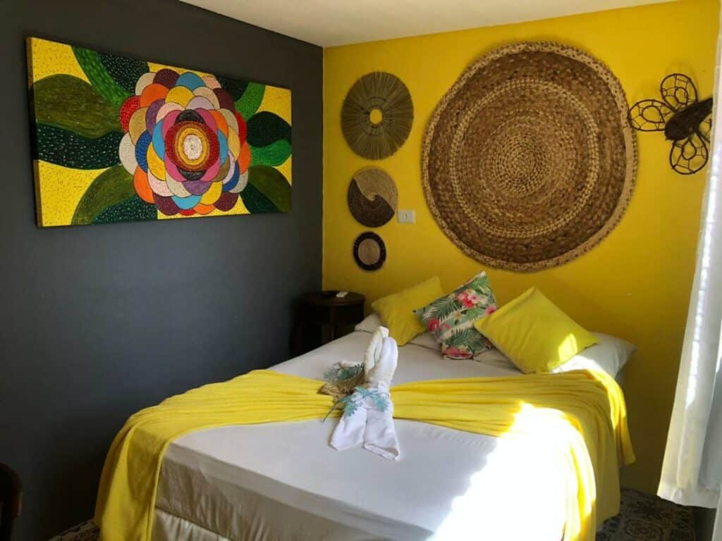 Quarto na Ladeira de Olinda com uma cama de casal decorada em tons de amarelo, com almofadas, as paredes do cômoda são amarelas e cinzas, com objetos decorativos pendurados