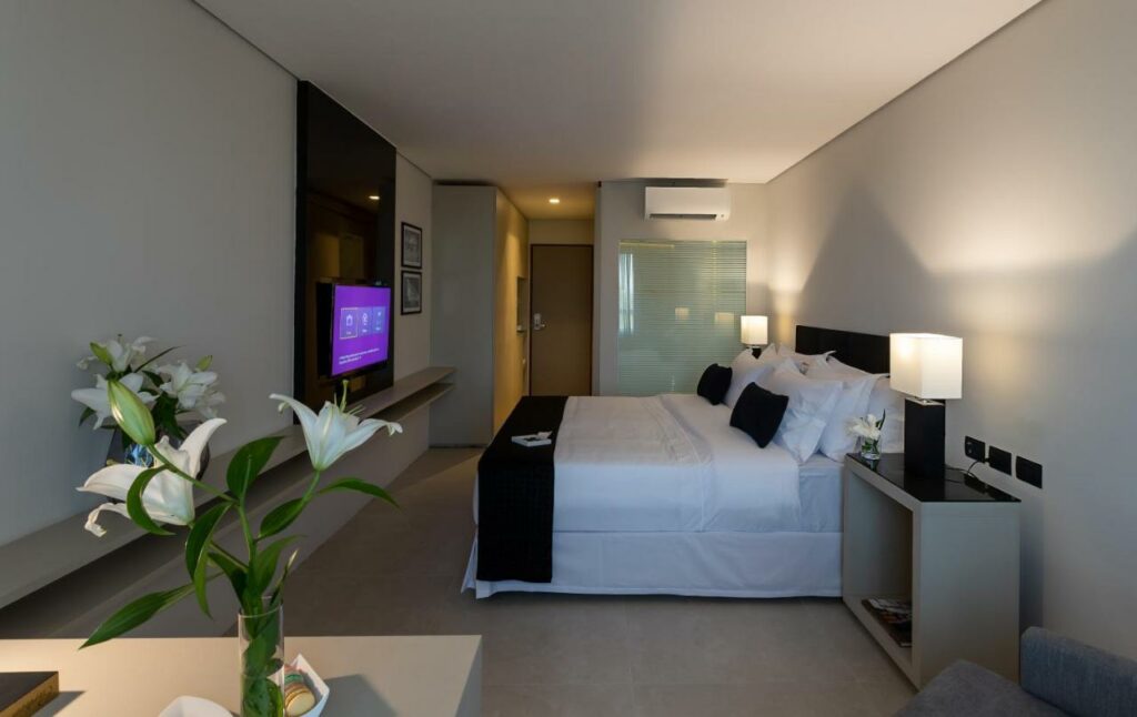 Quarto do LS Hotel com uma cama de casal, um abajur, ar-condicionado, uma televisão, itens de decoração minimalistas e alguns lírios brancos sob os móveis, para representar resorts em João Pessoa