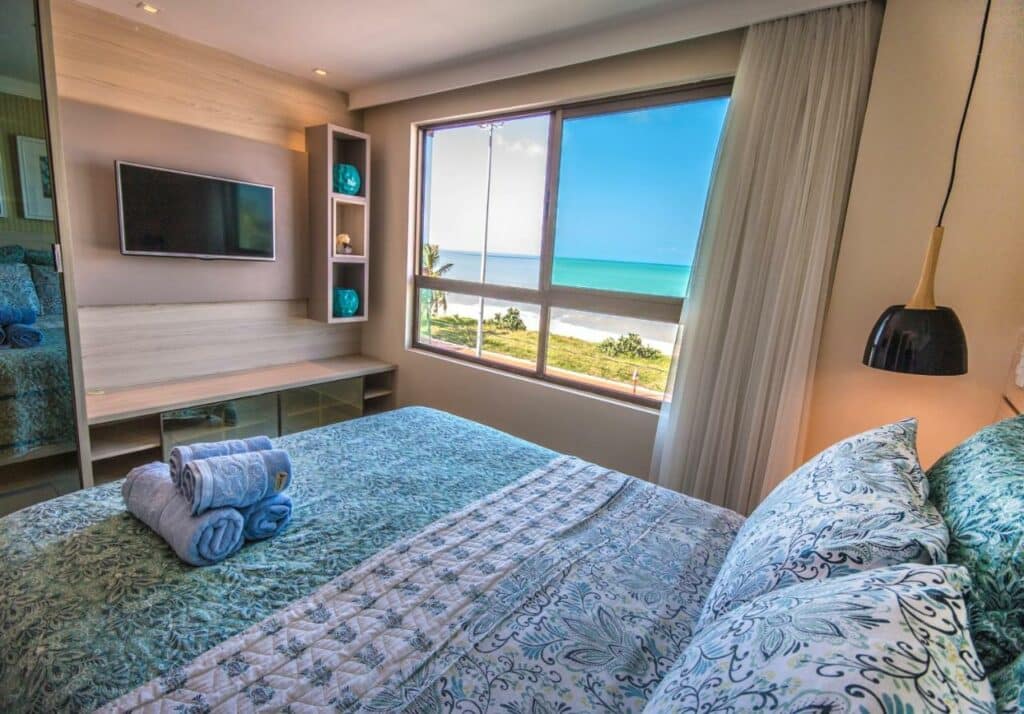 Quarto do Luxor Paulo Miranda Home Service com uma cama de casal, com uma janela ampla dando vista para a praia, uma televisão, uma luminária, para representar resorts em João Pessoa
