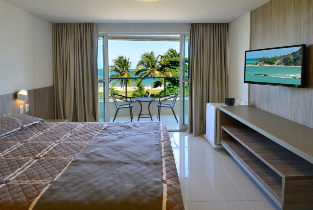 Quarto do Nord Luxxor Cabo Branco com uma varando dando vista para a praia, uma cama de casal, um frigobar e um rack, para representar resorts em João Pessoa