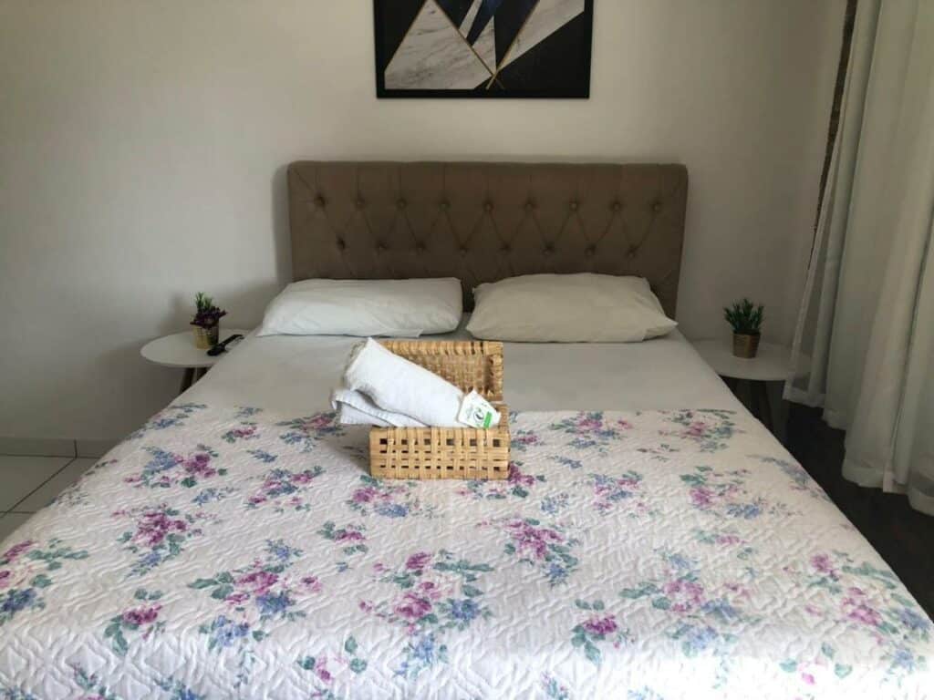 Quarto na Olinda mar pousada com uma cama de casal, dois travesseiros, uma coberta florida, uma caixinha com toalhas dentro, dois vasinhos de flores e um quadro