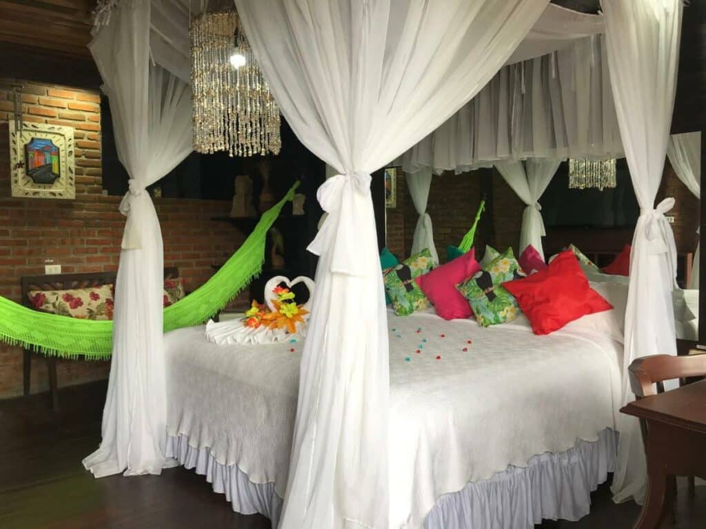 Quarto da Pousada Alto Astral  com cama de casal estilo bangalô com muitas cortinas marradas, a cama decorada com almofadas coloridas, um lustre no teto, ao lado e uma rede