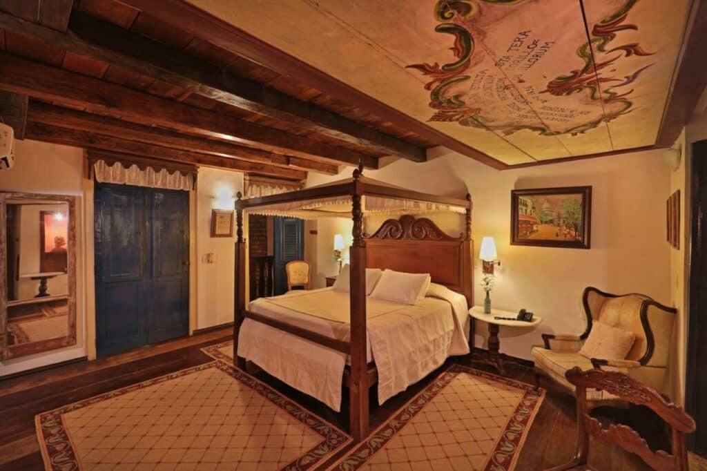 Quarto na Pousada do Amparo com uma cama de casal em estilo colonial, poltrona, quadros, abajur, tudo decorado em madeira, local amplo