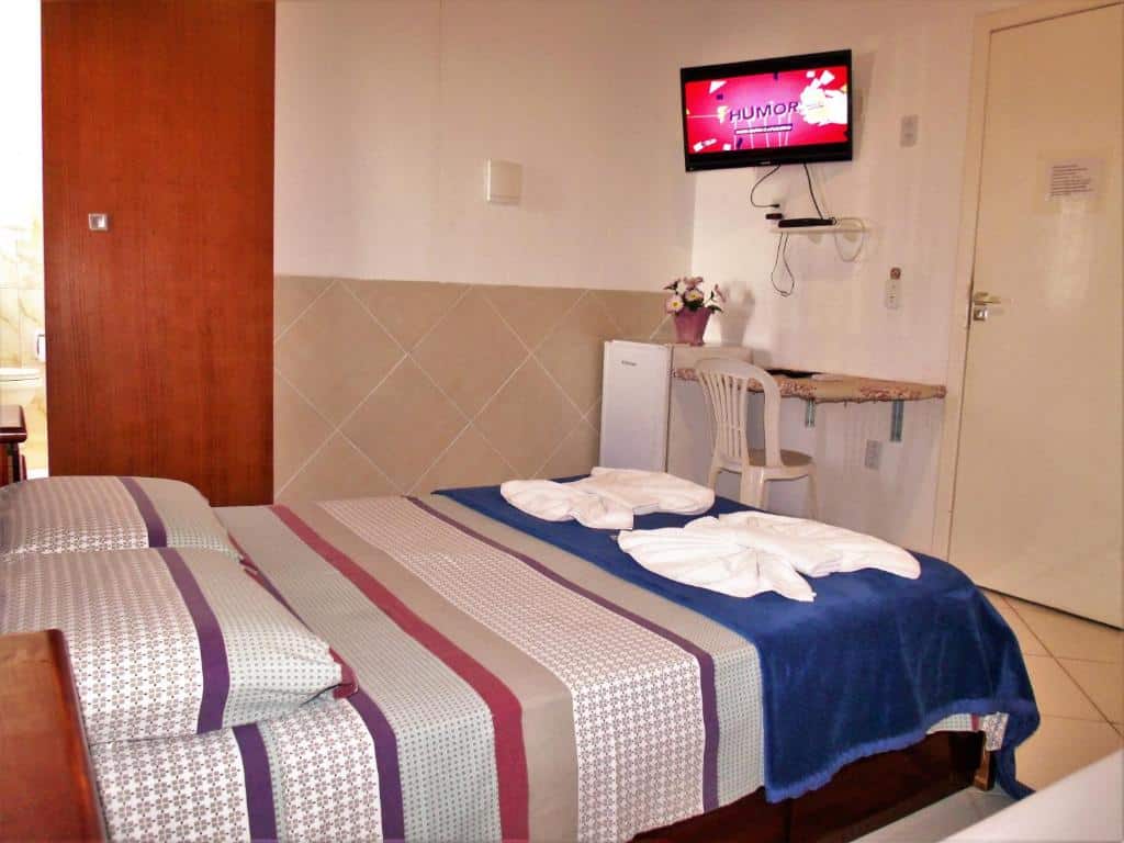 Quarto na Pousada Convento Da Penha com uma cama de casal, uma bancada com uma cadeira, uma televisão e um frigobar, para representar pousadas em Vila Velha