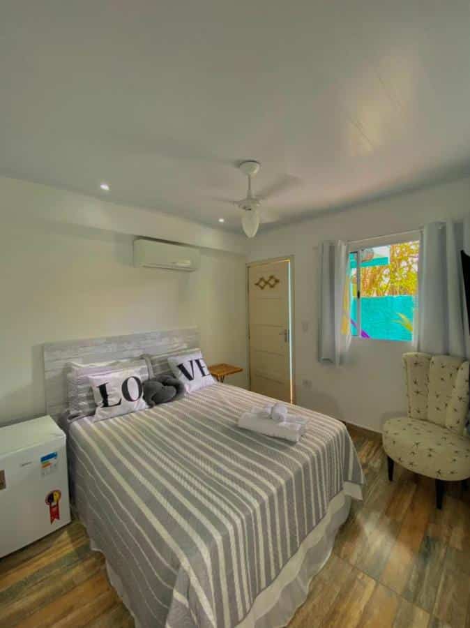 Quarto na Pousada Orquídeas do Guarujá com uma cama de casal, um frigobar, um ar-condicionado e uma poltrona, além da janela com cortinas