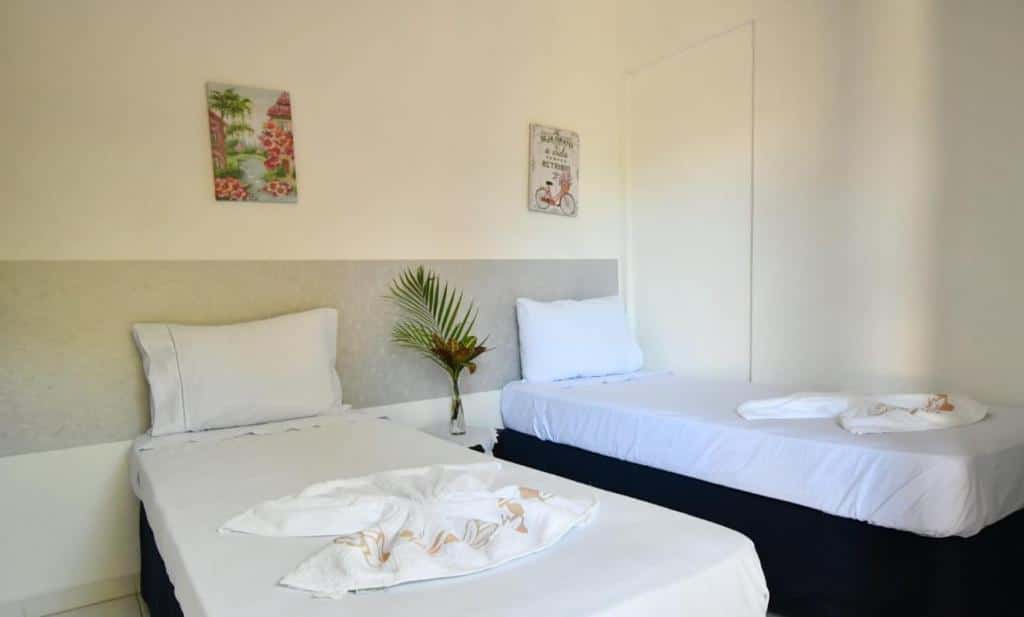 Quarto na Pousada Praia do Tombo com duas camas de solteiro, com toalhas e travesseiros, um vaso de flor e alguns pequenos quadros na parede