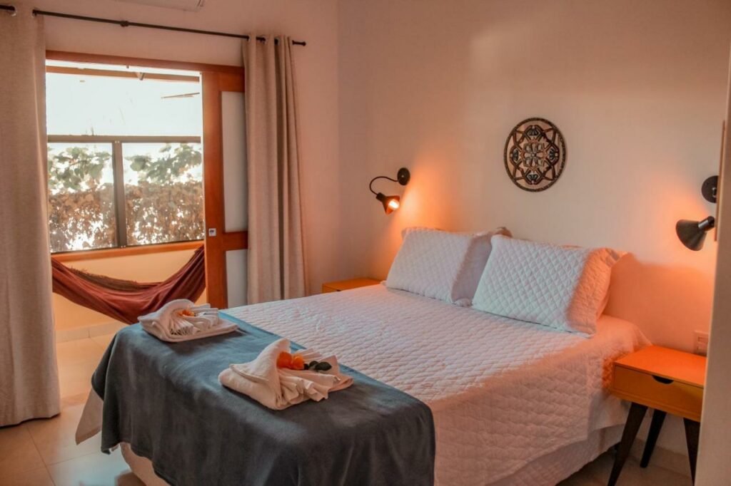 Uma cama bem arrumada da Pousada Quintal da Lua com roupa de cama branca, mesas de cabeceira, luminárias de parede e uma rede perto de uma janela com cortinas fechadas.