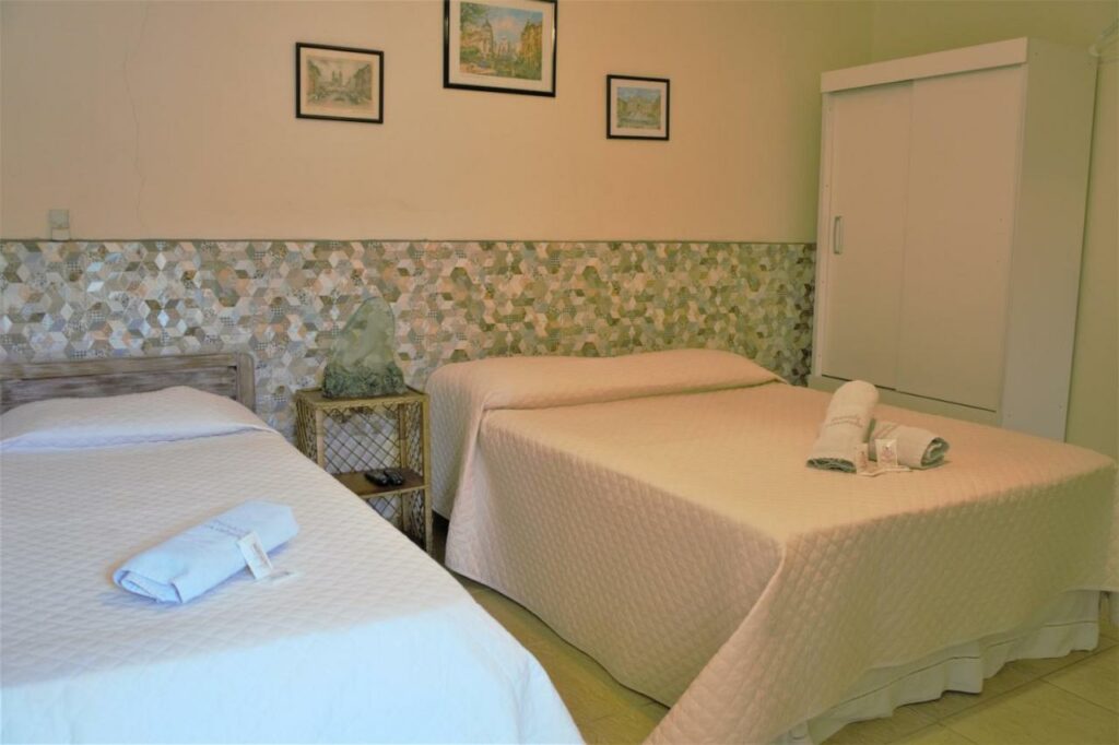 Quarto na Pousada Sorocotuba com uma cama de casal e uma de solteiro, um pequeno armário, decoração em tons de verde claro e branco, toalhas e travesseiros sob a cama