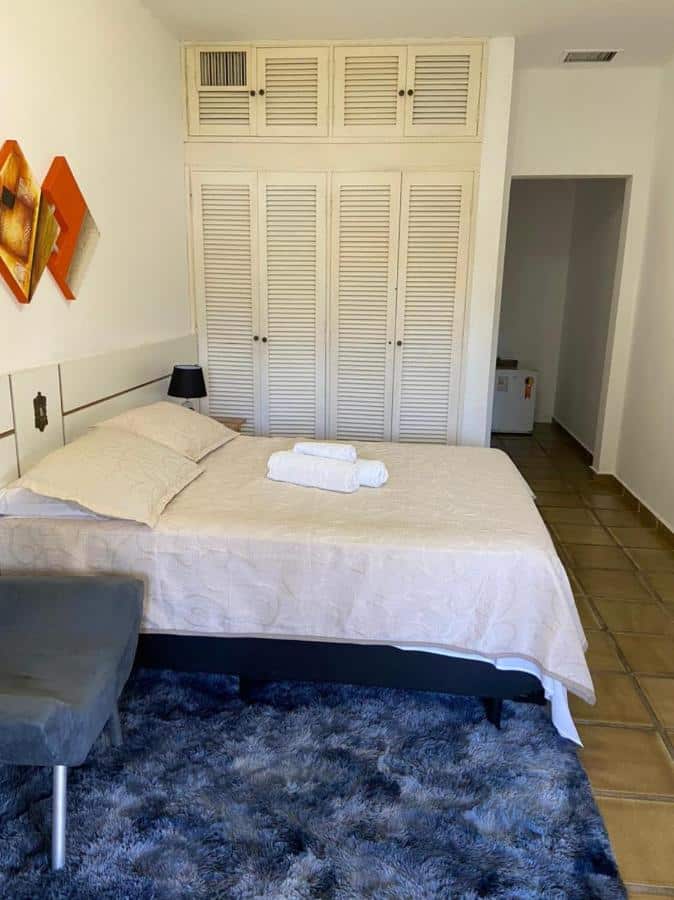 Quarto na Pousada Vila Novo Tempo com cama de casal, um armário, uma poltrona e um tapete, tudo em tons de branco e azul