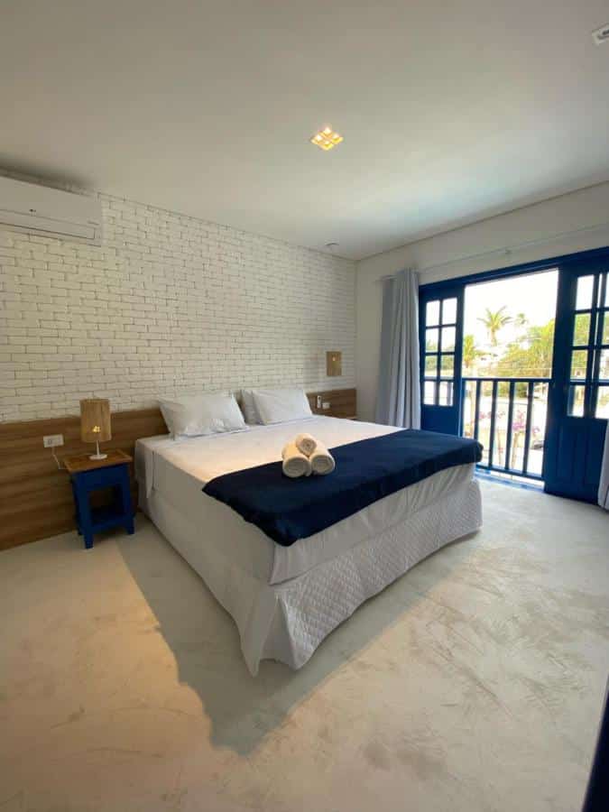 Quarto da Pousada Vila Santô com tudo decorado em branco e azul, uma cama de casal com travesseiros e toalhas, uma ampla sacada, um ar-condicionado e dois abajures