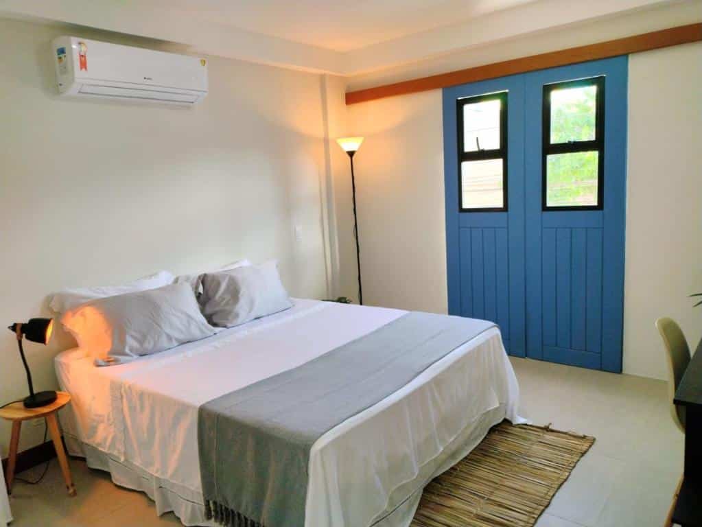 Quarto na Pousada VilaZinha tudo decorado em tons de azul e branco, uma cama de casal, dois abajures, travesseiros sob a cama e um tapete de bambu falso