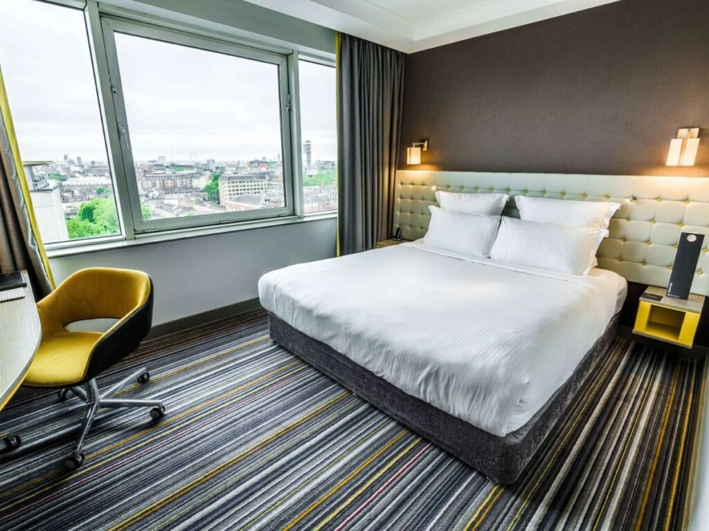 Quarto do Pullman London St Pancras com uma ampla janela dando vista para a cidade, uma cama de casal, iluminação indireta na cabeceira da cama e uma mesa de trabalho com cadeira, para representar hotéis em Londres