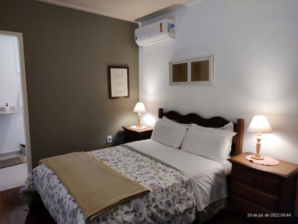 Quarto no Rancho São Carlos com uma cama de casal, dois abajures, um ar-condicionado, local decorado e novo, com travesseiros e cobertas sob a cama