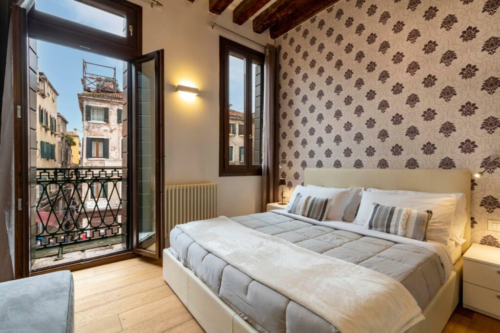 Quarto no Residence Le Beccarie - Rialto com uma varanda virada para o centro, uma janela, ar-condicionado, uma cama de casal, chão de madeira, tudo em tons de branco e cinza