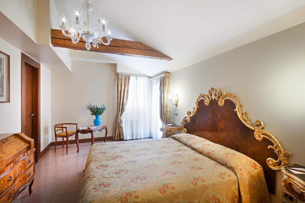 Quarto amplo no Residenza d'Epoca San Cassiano, com um lustre, duas janelas, uma mesa com uma cadeira, uma cômoda, uma cama de casal ampla, tudo decorado em madeira e com detalhes em dourado