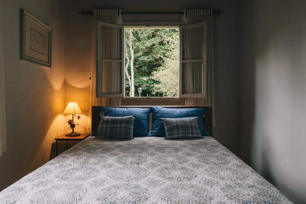 Quarto no Sítio Valfenda Itatiaia com uma cama de casal com almofadas e travesseiros, uma janela com vista para a natureza, um abajur e uma quadro