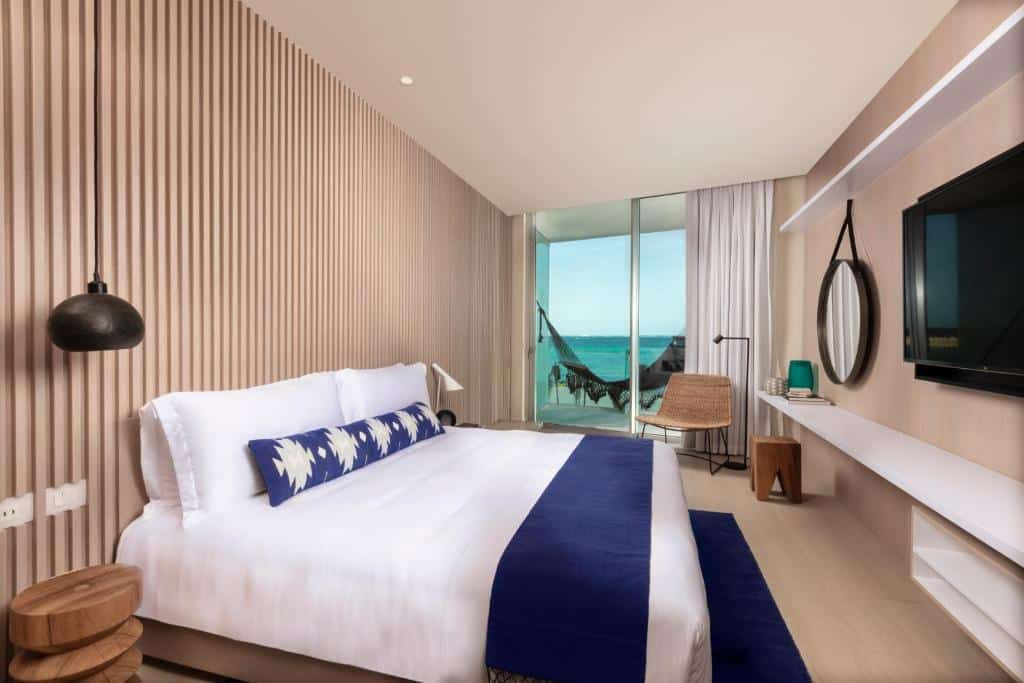 Quarto no SLS Cancun Hotel & Spa com uma cama de casal, uma sacada virada para o mar com uma rede pendurado, um espelho, uma televisão, uma poltrona e dois abajures, local decorado em tons de branco e azul, para representar hotéis em Cancun