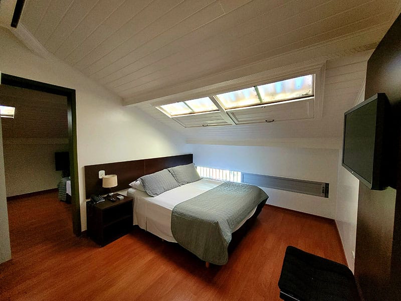 quarto de teto inclinado com uma claraboia em cima da cama de casal, e com uma porta que dá acesso a outro quarto com cama de casal