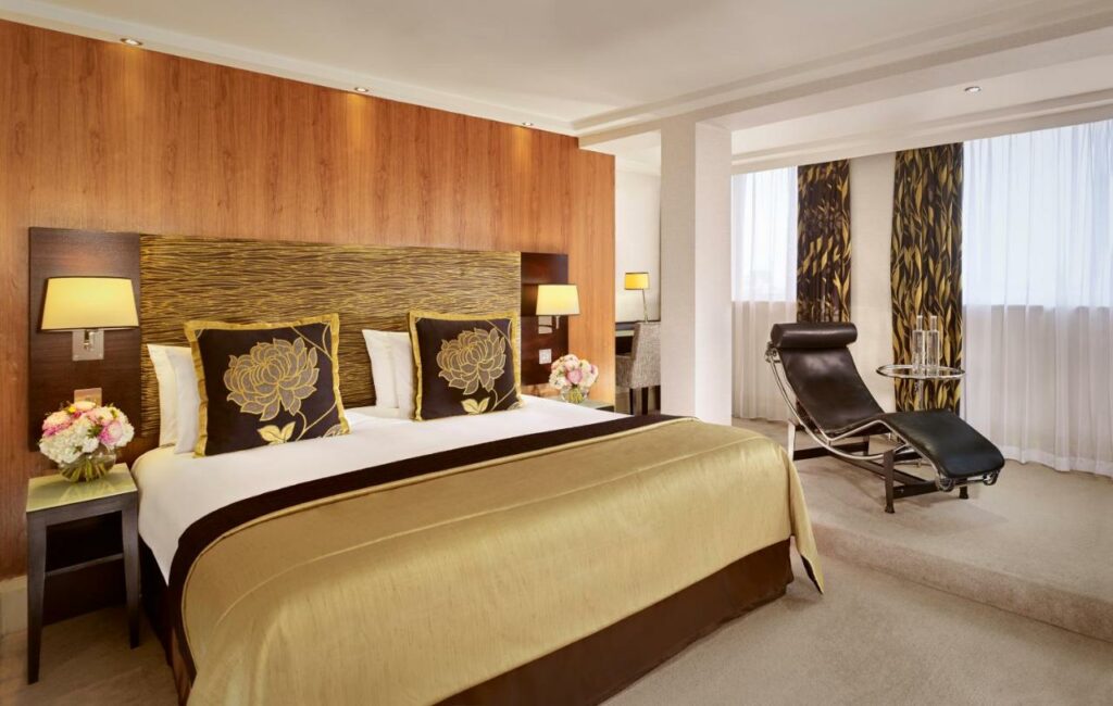 Quarto no The Cavendish London, local amplo, com duas janelas, uma cadeira estilo espreguiçadeira, uma cama de casal, com almofadas e travesseiros, dois abajures nas mesinhas de cabeceira, tudo decorado em tons de marrom e dourado, para representar hotéis em Londres