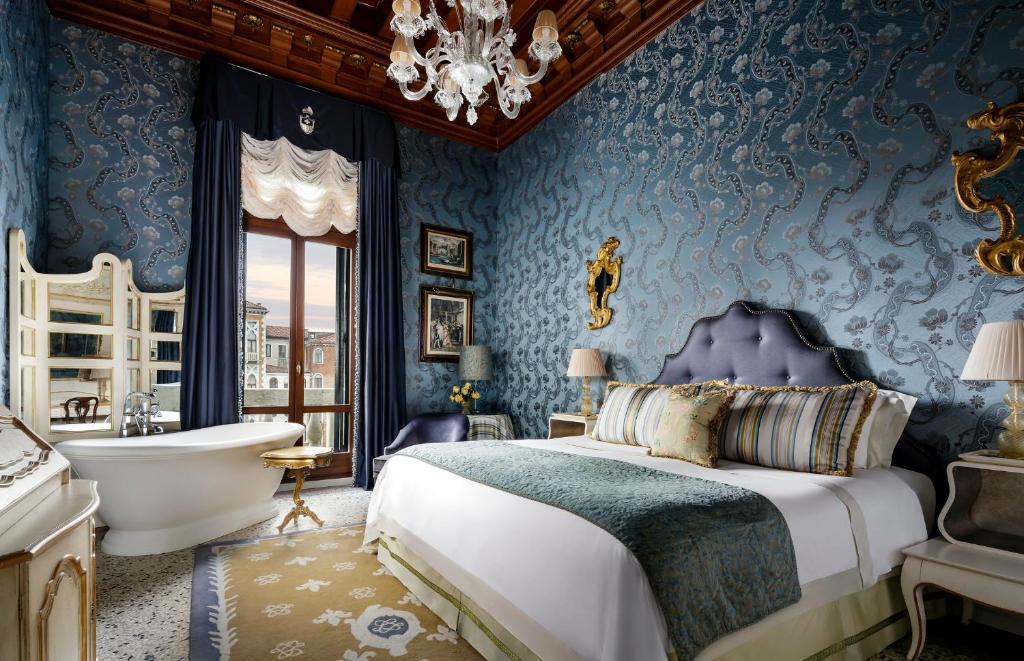 Quarto do The Gritti Palace ambiente muito amplo, com uma sacada, uma banheira, uma poltrona, uma cama de casal, uma cômoda, uma mesinha de cabeceira com um abajur, o local é inteiro decorado em tons de dourado e azul, tudo muito elegante e há um lustre no teto, para representar hotéis em Veneza