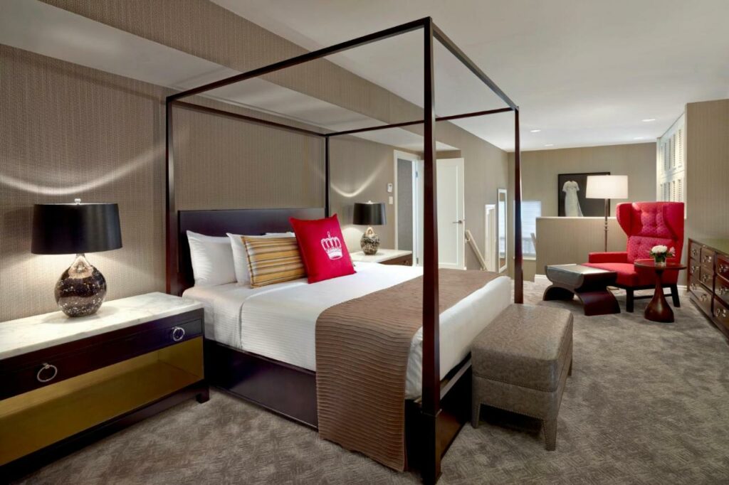 Quarto no The Omni King Edward Hotel com uma cama de casal, uma enorme poltrona, uma cômoda com um abajur, chão de carpete e tudo decorado em tons de marrom, bege e vermelho