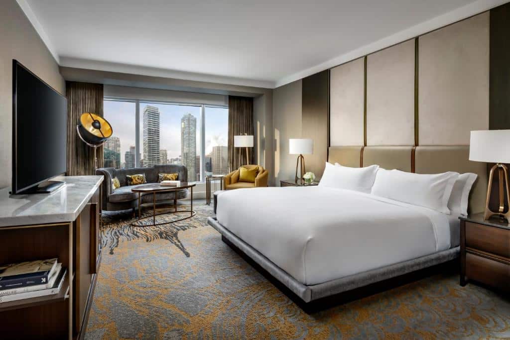Quarto amplo no The Ritz-Carlton com uma enorme janela com vista para a cidade, um pequeno sofá com almofadas, uma poltronas, três abajures, uma televisão, uma cama de casal e um carpete em tons de azul e dourado, para representar hotéis em Toronto