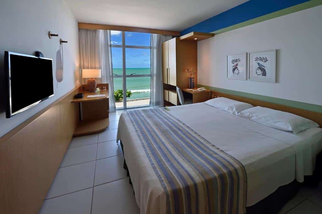 Quarto do Verdegreen Hotel com uma cama de casal, uma sacada com vista para o mar, uma televisão, uma pequeno armário e um abajur