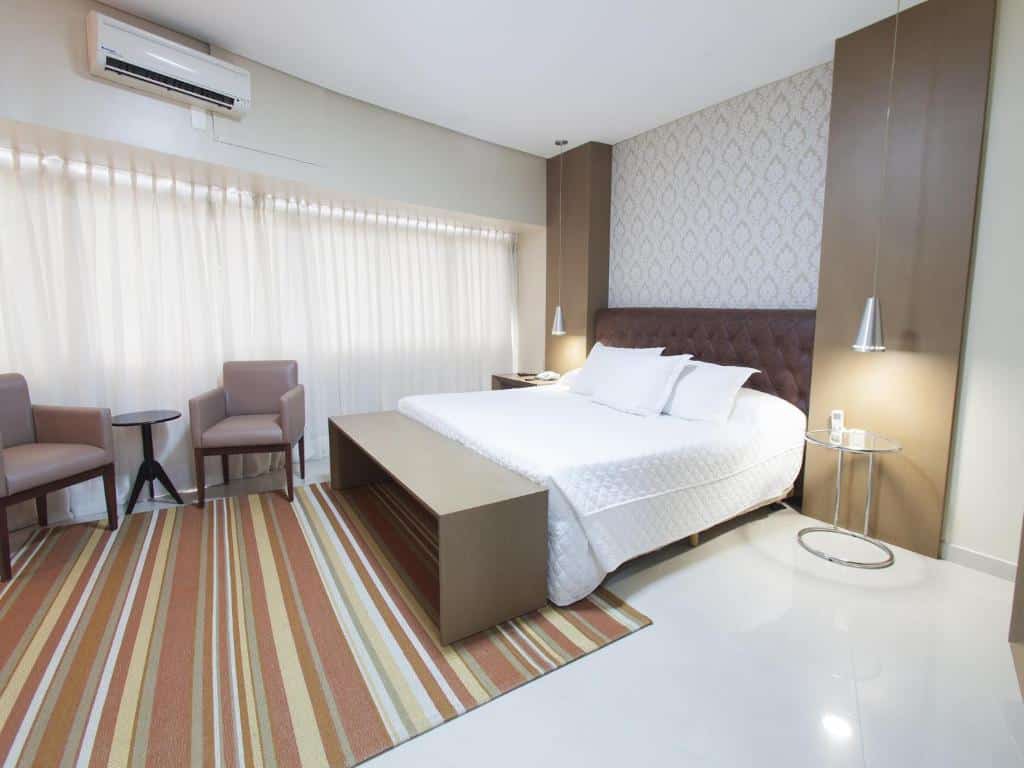 Quarto do Hotel Village Premium Joao Pessoa com uma cama de casal, tudo decorado em tons de marrom, branco e bege, uma pequena mesinha com duas poltronas, um abajur, uma sacada com cortinas, para representar resorts em João Pessoa