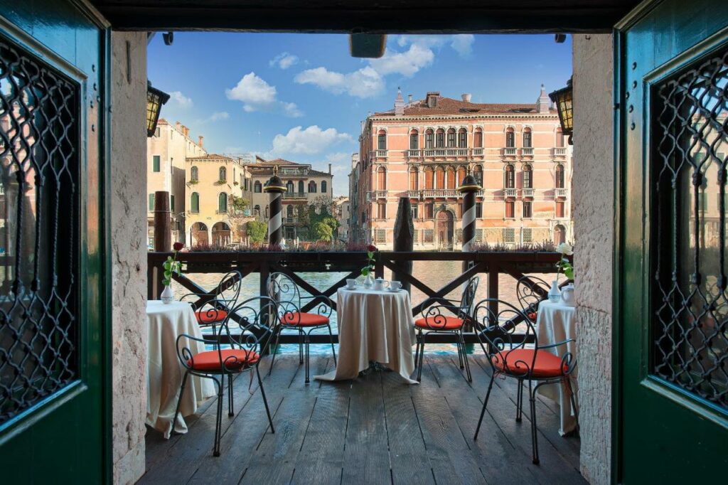 Terraço do Residenza d'Epoca San Cassiano com mesas redondas e cadeiras vermelhas dando vista para o canal de Veneza, para representar hotéis em Veneza