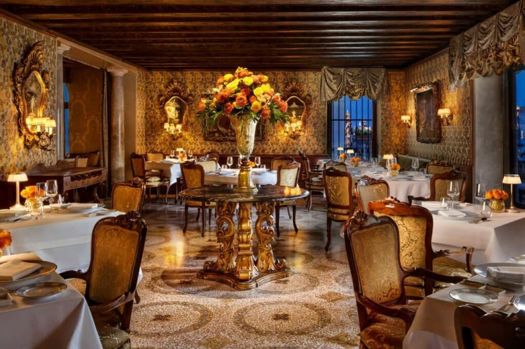 Restaurante do The Gritti Palace inteiro decorado em estilo renascentista, com papéis de parede, muitas cortinas, quadros, espelhos e abajures nas paredes, no centro do local há uma mesa de mármore com um enorme vaso de flores, ao redor, estão as mesas e cadeiras decoradas com flores e pequenas luminárias