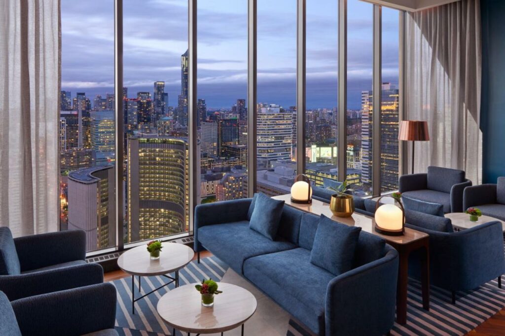 Vista do lounge compartilhado no Sheraton Centre Toronto Hotel com uma janela de vidro ampla ao lado dando vista para a cidade, com poltronas azuis e algumas mesinhas de centro
