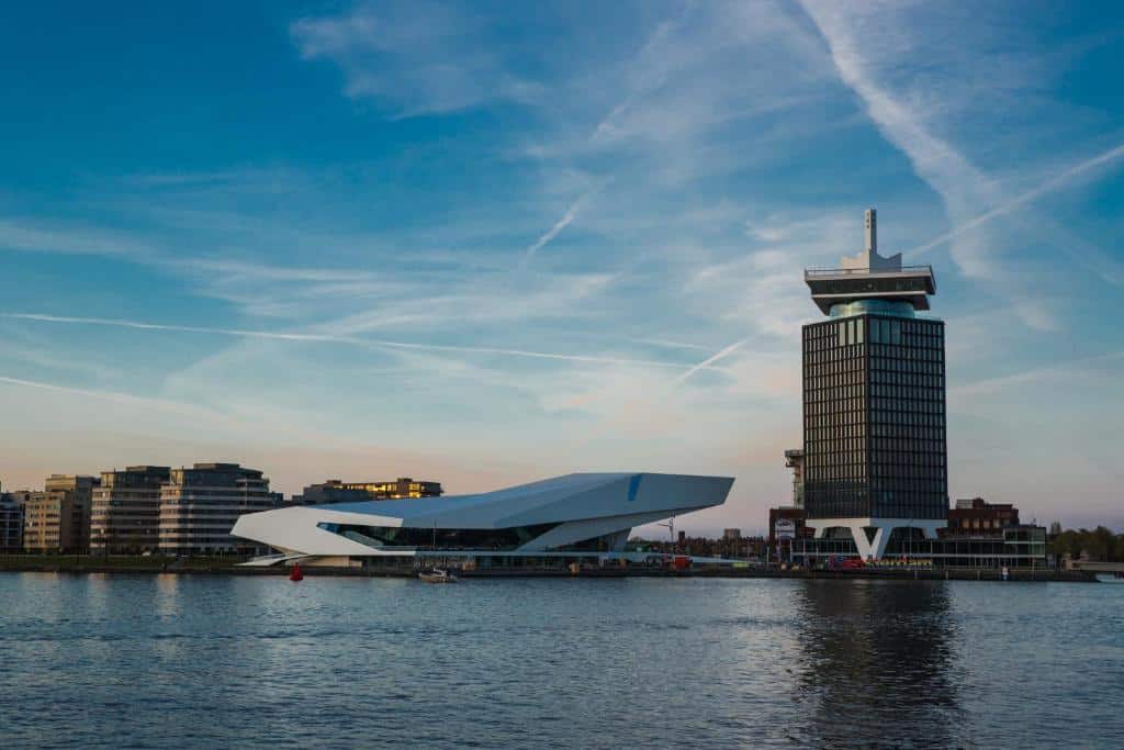 Rio com o prédio do Sir Adam Hotel, em Amsterdam, com um edifício ao lado num formato oval horizontal e ao fundo tem o céu com poucas núvens