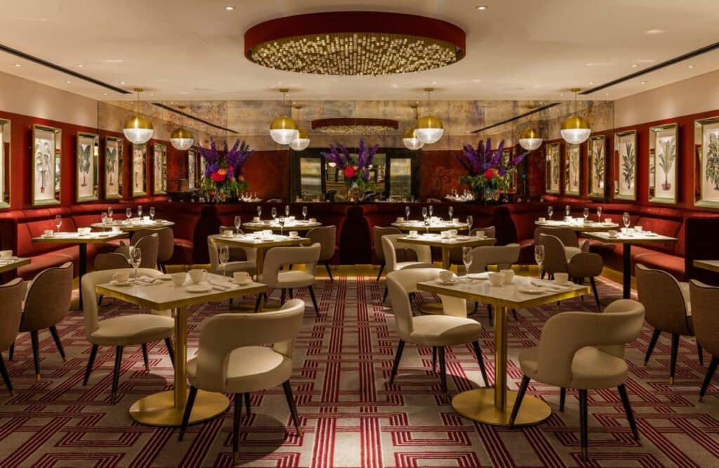Restaurante do Strand Palace Hotel decorado em tons de dourado e vermelho, com vasos de flores e ambiente elegante