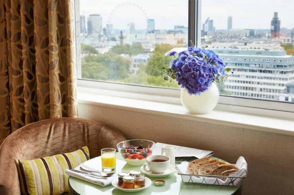 Janela do quarto do The Cavendish London com vista para a cidade, com um vaso de flor, uma pequena mesa e duas poltronas