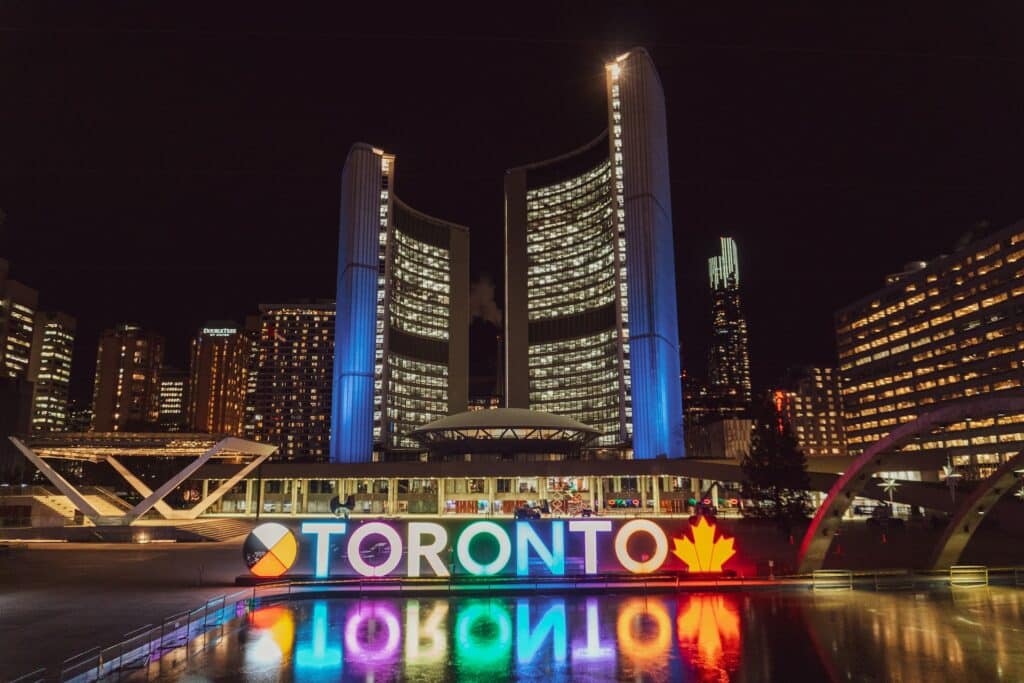 Vista dos prédios iluminados em Toronto, com uma placa colorida escrito o nome da cidade com um lago na frente