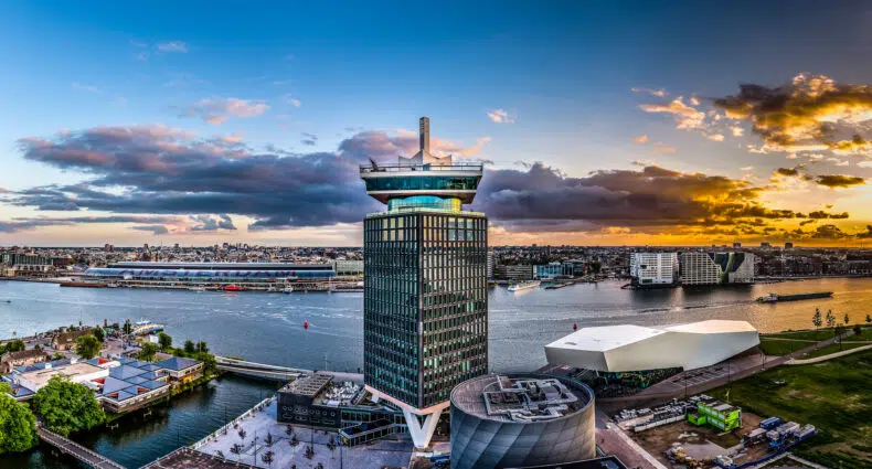 Vista da Torre A'DAM, onde fica o Sir Adam Hotel, um edifício alto e moderno com deck de observação em um rio, cercado por outros edifícios, durante o pôr do sol com nuvens no céu. Ilustra o post de hotéis em Amsterdam.