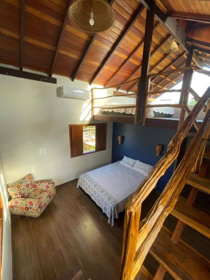 Chalé na Vila Ypáwa com dois andares, com uma cama de casal, uma poltrona e uma janela, do lado direito, uma escada de madeira leva para o mezanino, para representar pousadas em Alter do Chão