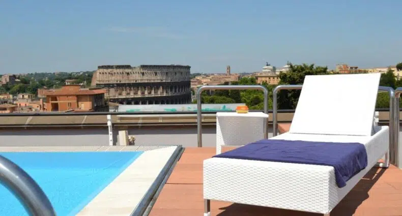 Uma espreguiçadeira branca ao lado de uma piscina em um telhado com o Coliseu visível ao fundo sob um céu claro, no Mercure Roma Centro Colosseo, para representar hotéis em Roma.