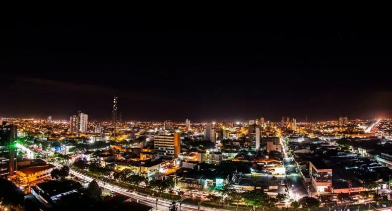 Uma paisagem urbana à noite com edifícios iluminados, ruas movimentadas e luzes brilhantes sob um céu escuro.