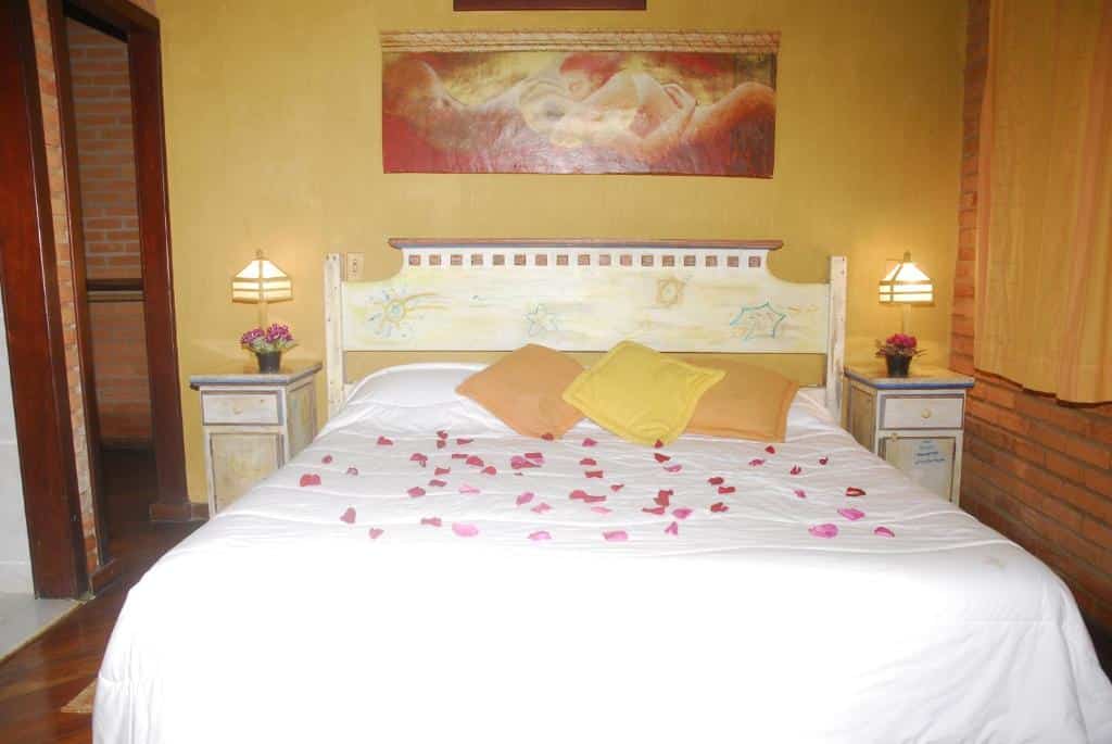 Quarto no Vista Linda Hotel com uma ampla cama de casal com almofadas e pétalas de rosa, duas mesinhas de cabeceira com abajures