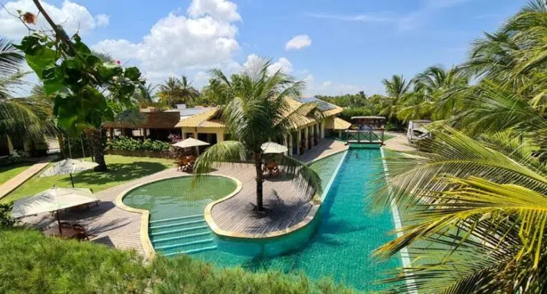 Uma grande piscina externa com uma ilha de palmeiras, espreguiçadeiras, guarda-sóis e paisagismo tropical sob um céu azul, na Pousada Coqueiro Beach, para representar pousadas em Luís Correia.