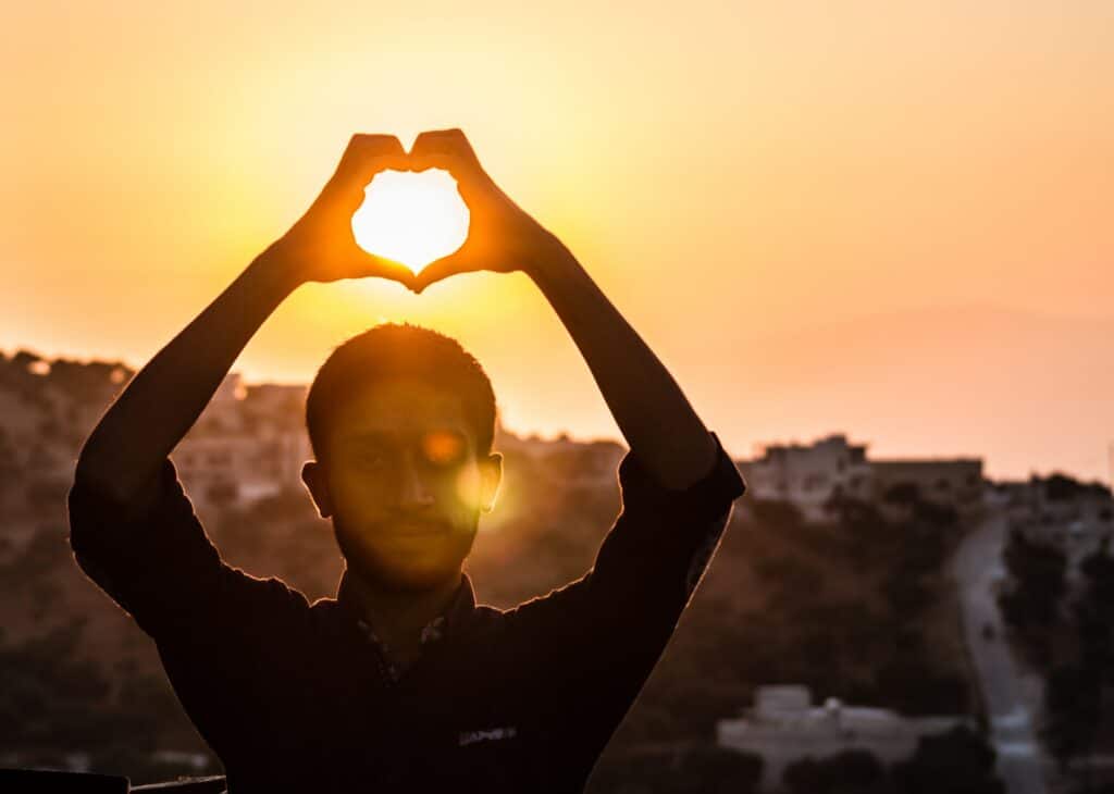 Homem fazendo coração com as mãos, com um pôr do sol amarelado atrás, no Aaqrabâte, vilarejo localizado na Síria