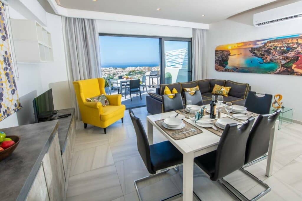 Sala de estar do Artist Terrace Apartments com uma ampla varanda com vista para a cidade e o mar, o local conta com uma mesa de seis lugares, um sofá, uma poltrona, uma televisão, tudo em tons de preto, cinza, branco e amarelo