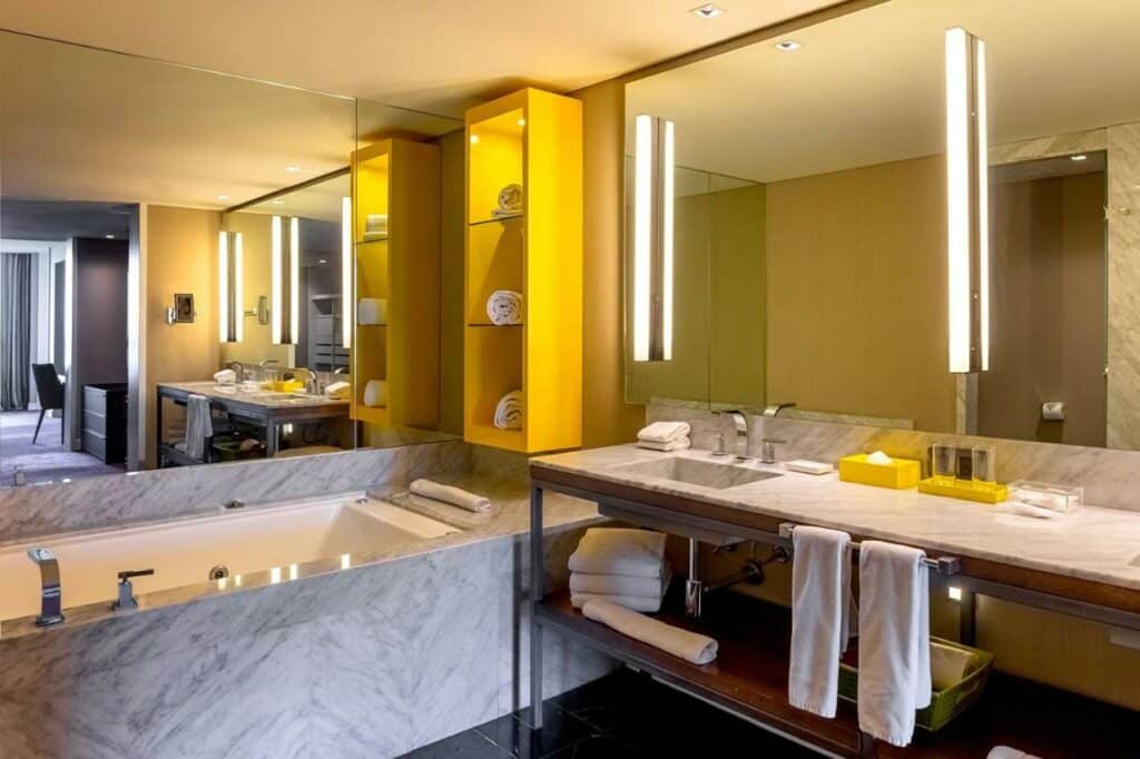 Banheiro no Hyatt Centric Montevideo com uma banheira, duas pias, muitos espelhos e armários de conceito aberto com toalhas, tudo com detalhes em amarelo
