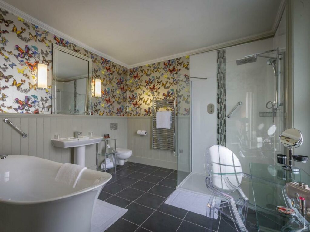Banheiro amplo do The Fleet com uma banheira, box de vidro, pia, espelho, vaso sanitário e uma penteadeira, há um papel de parede com desenho de borboletas em uma parte da acomodação