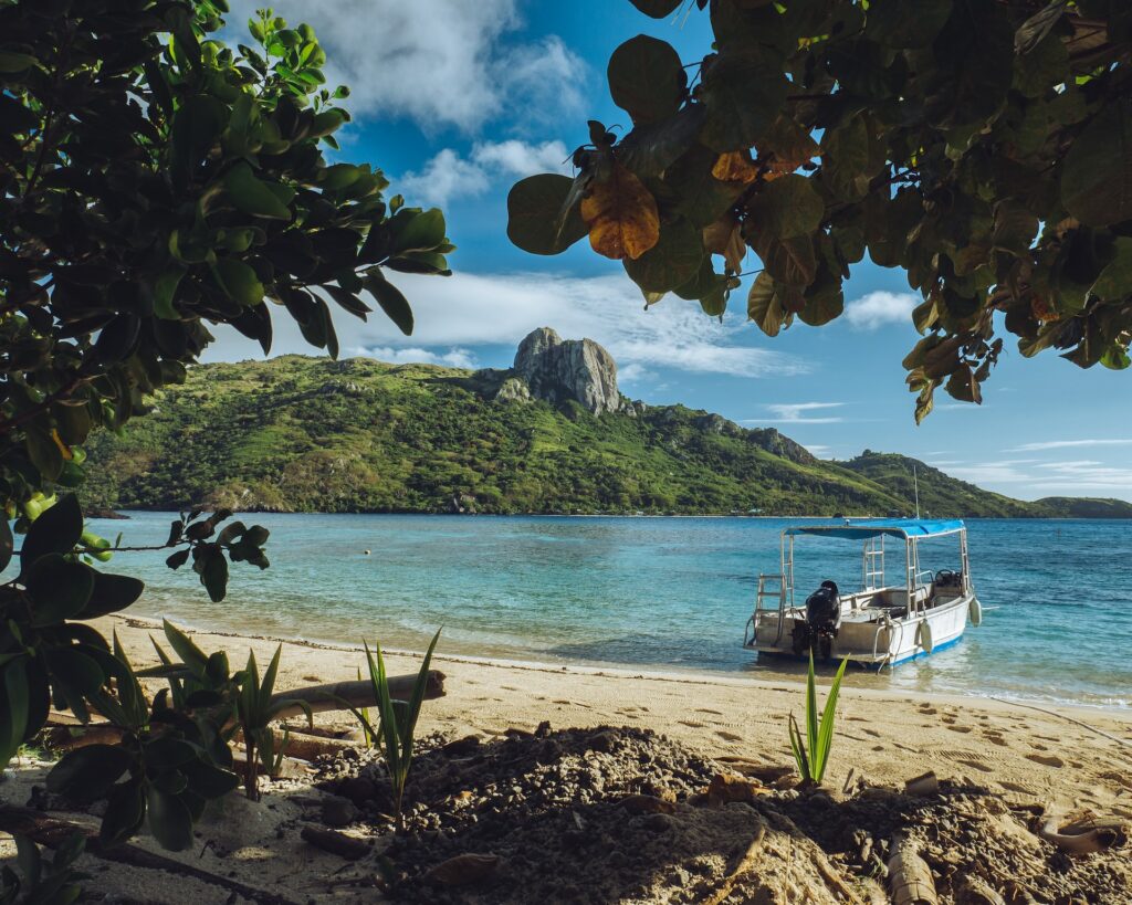 Barco azul e branco ancorado na areia de uma praia em Fiji para ilustrar o seguro viagem Fiji. Há vegetação ao redor e a água é cristalina. - Foto: Nicolas Weldingh via Unsplash
