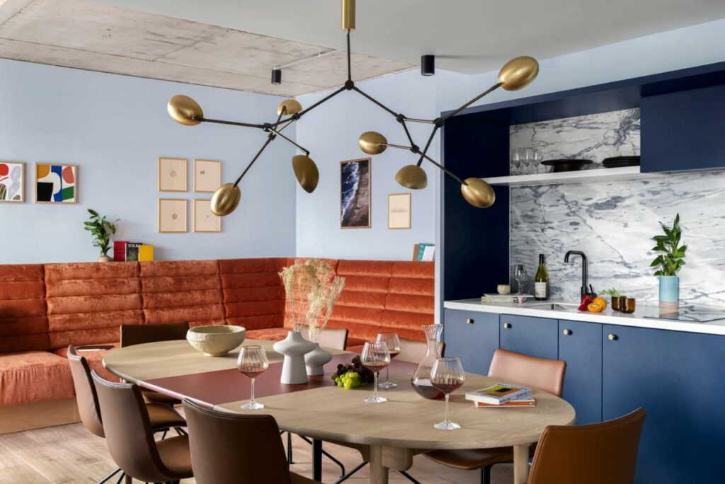 Sala de jantar no Beckett Locke com uma mesa de oito lugares, alguns sofás, uma luminária cheia de lâmpadas, tudo decorado em tons de azul, madeira, cobre e laranja