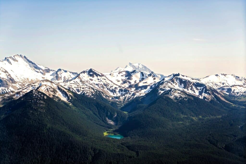 Conjunto de montanhas glaciais com neve em seus cumes, cercado por natureza e um pequeno lago com águas azuis cristalinas ao meio para representar o seguro viagem Whistler.