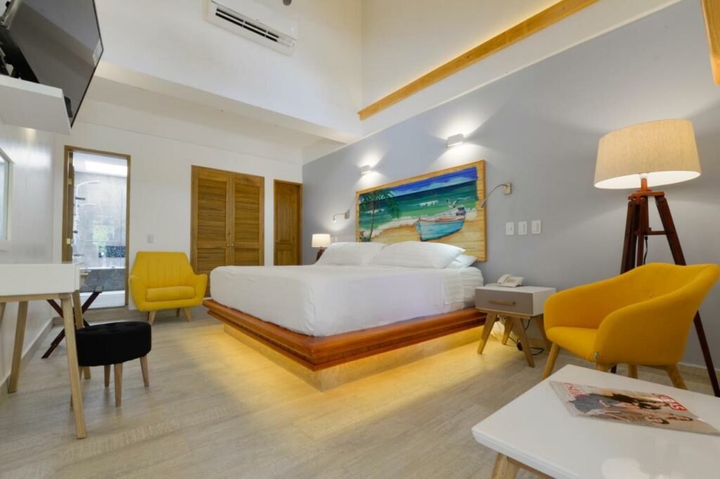Quarto amplo na Casa Las Palmas Hotel Boutique com uma cama de casal, duas poltronas amarelas, um abajur de chão, uma mesa de escritório e um armário embutido, tudo em tons de amarelo, madeira e branco
