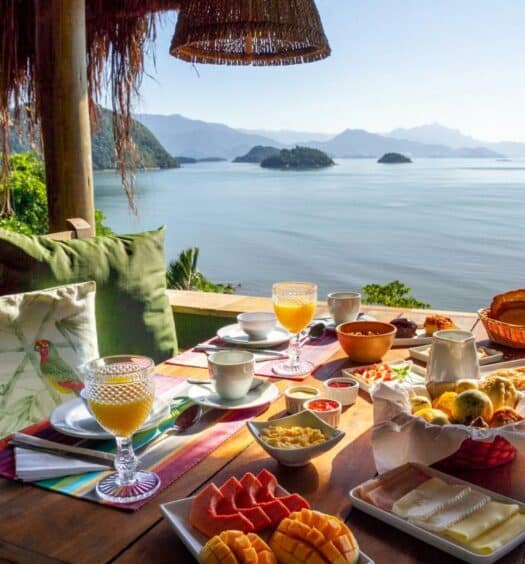 Varanda da Casa Luz com uma mesa com um café da manhã e o local dando vista para o mar e as montanhas
